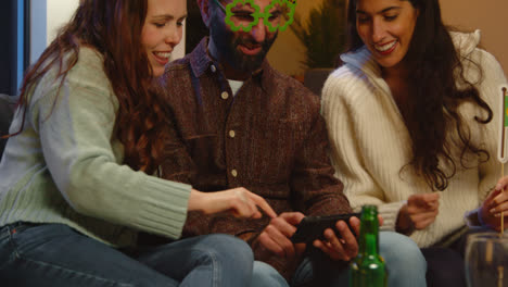 Group-Of-Friends-Dressing-Up-At-Home-Or-In-Bar-Celebrating-At-St-Patrick's-Day-Party-Looking-At-Photos-On-Phone-2